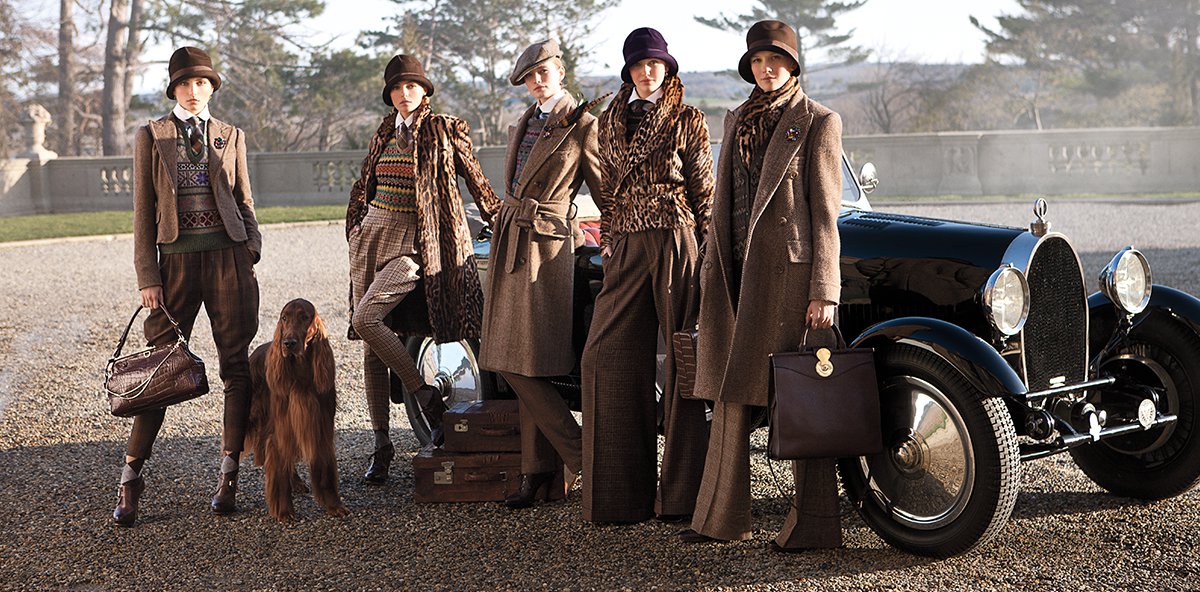 Models wear various brown outfits posing in front of old-fashioned car.