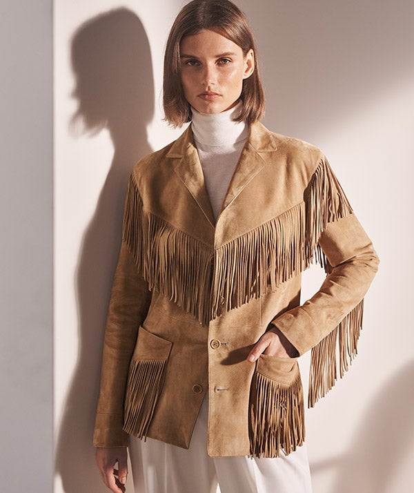 Woman in tan fringe jacket & off-white turtleneck & trousers