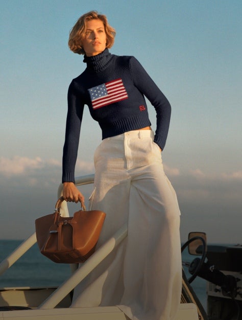 Woman holding brown leather bag wears navy sweater with American flag and white wide-leg trousers.