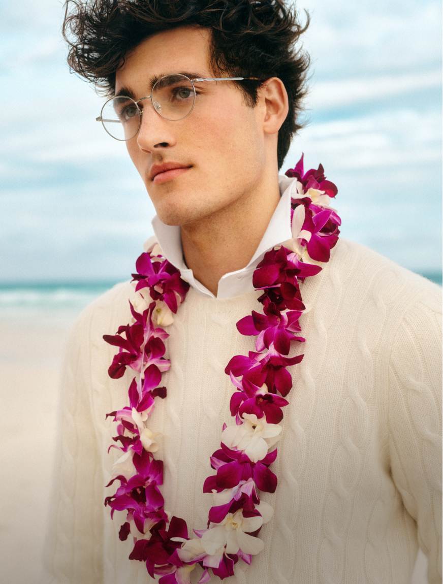 Man at beach wears a white cable-knit cashmere sweater and a purple lei.