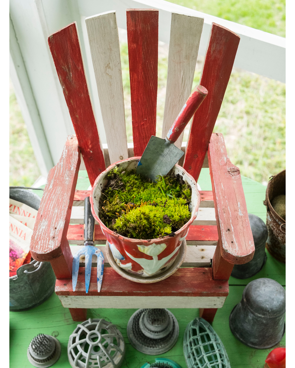 A secret garden of moss nestled in a vintage flowerpot takes a seat in a doll-sized Adirondack chair surrounded by a collection of flower arrangement frogs