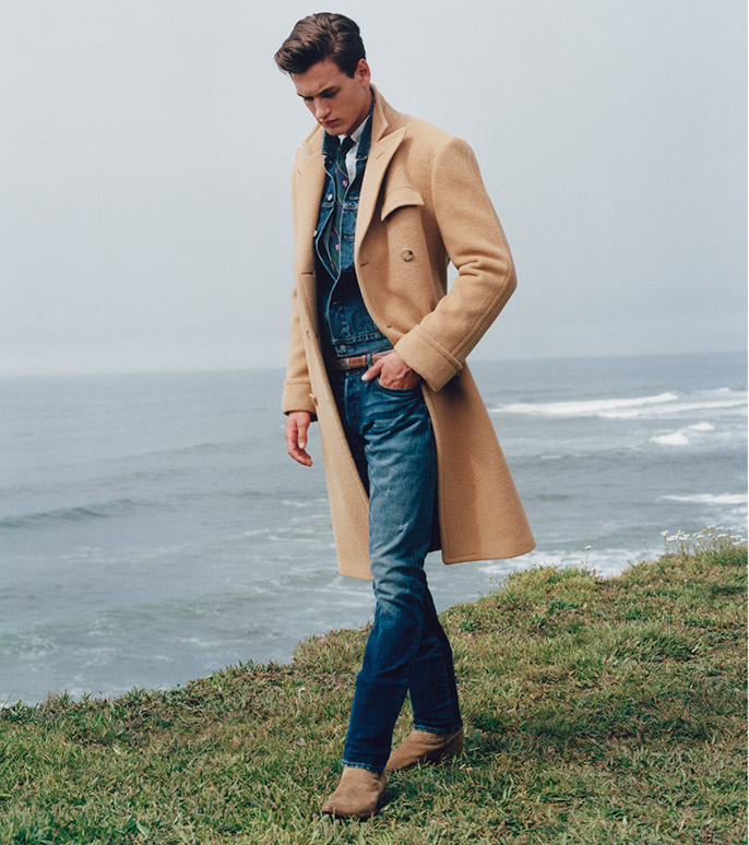 Man by water in tan peacoat layered over denim jacket & jeans 