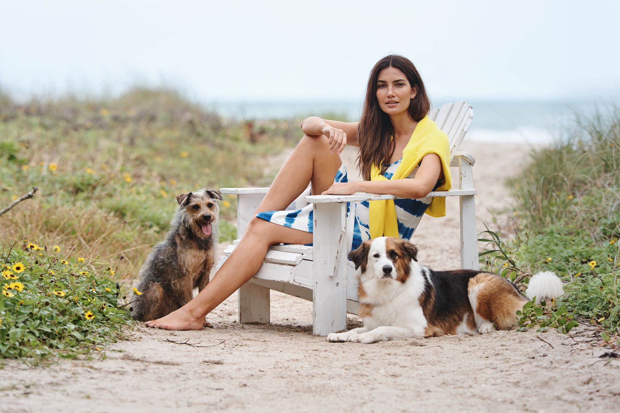 Woman in tie-dye dress sits on chair on beach with dogs.