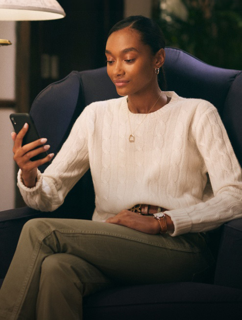 Woman in chair wearing white cable-knit sweater looks at cellphone.