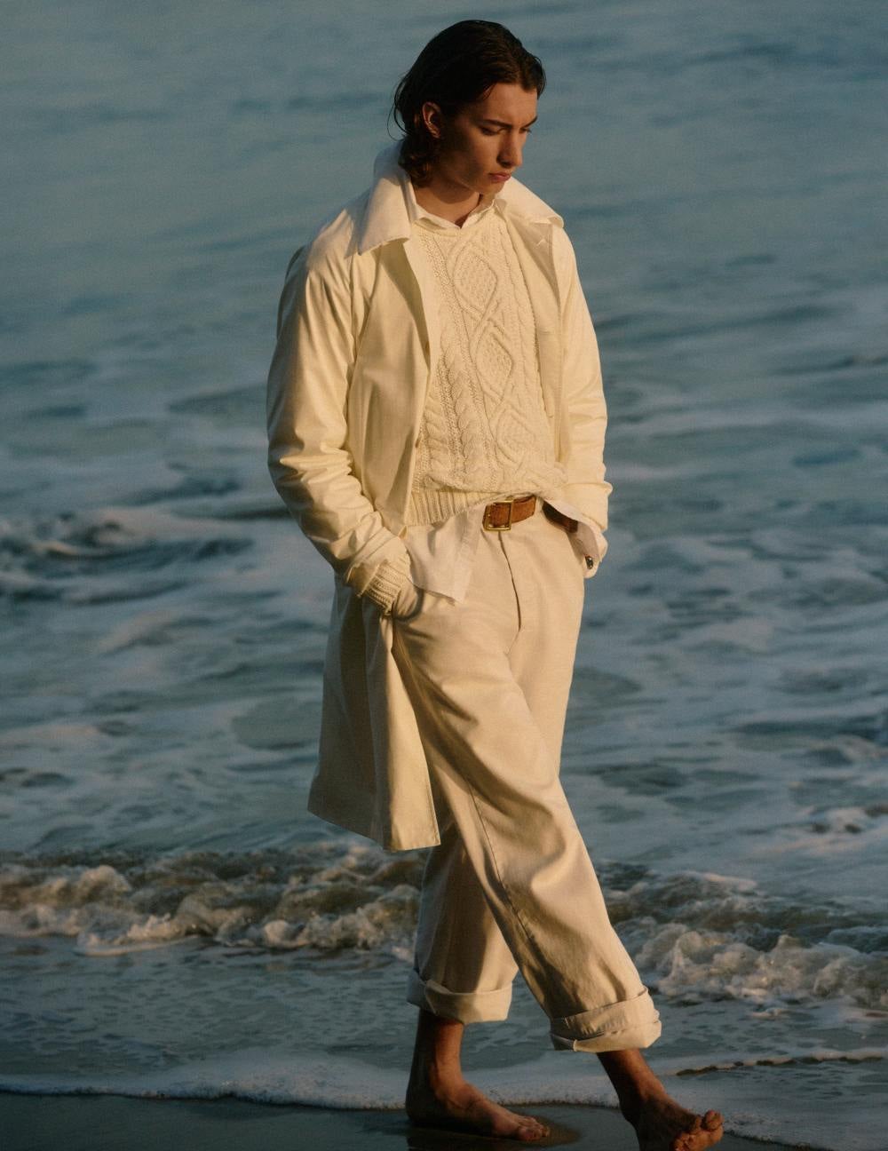 Man walking on beach wears cream trench, Aran-knit sweater, and chinos.