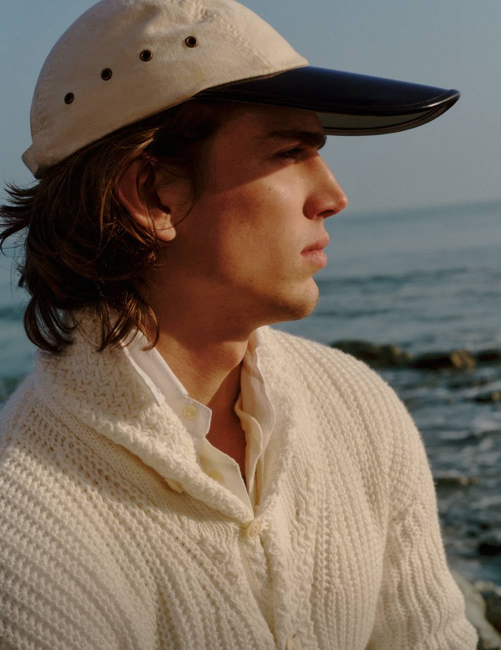 Man wears white knit cardigan and white ball cap with navy brim.