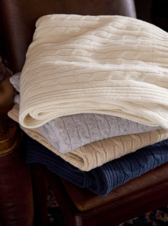 Folded stacked cable-knit blankets in cream, grey, tan, and navy.