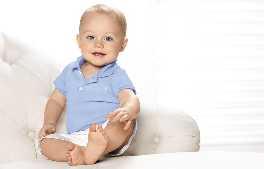 Baby in light blue Polo shirt & white short sits in white tufted armchair