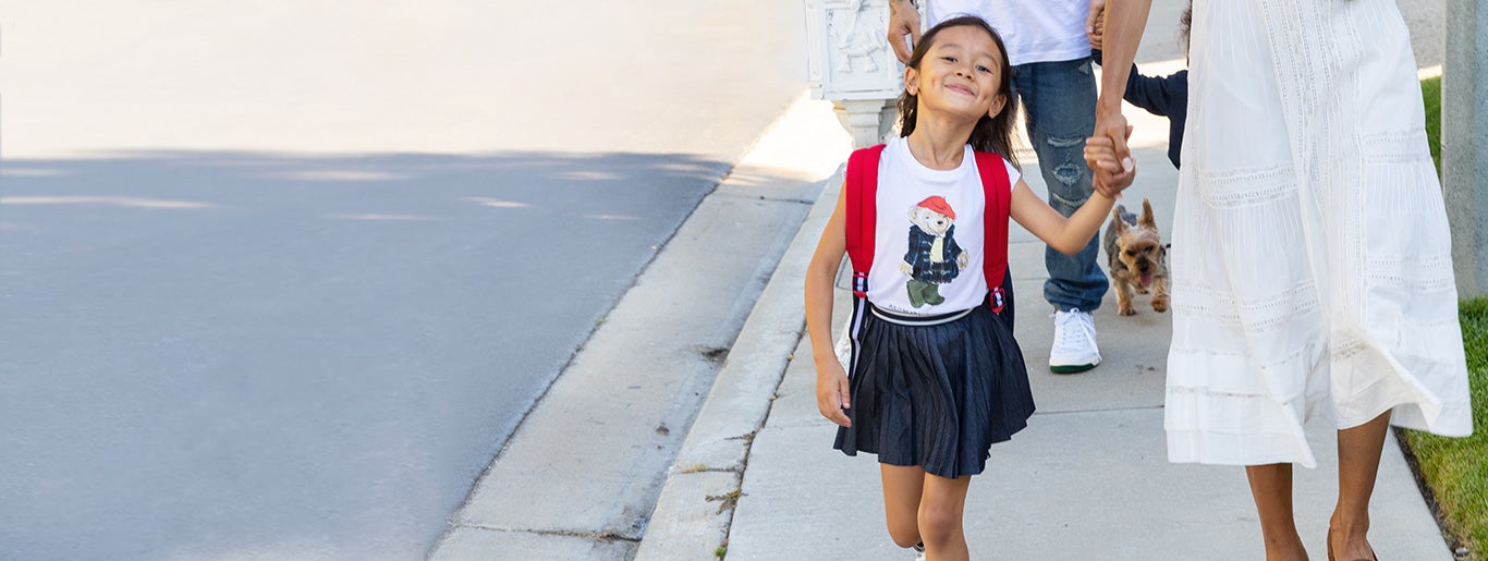 Girl wears Polo Bear T-shirt and pleated skirt while walking on sidewalk.