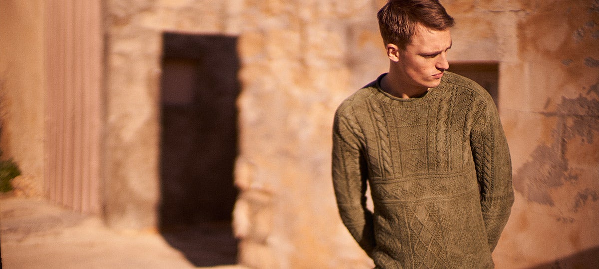 Man wears fisherman's sweater in front of stone building.